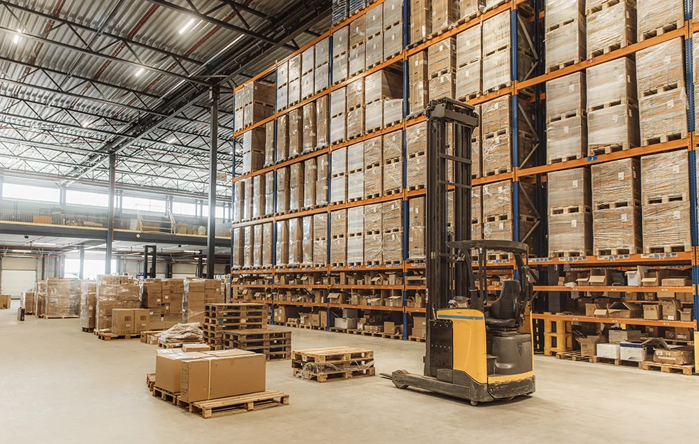 Large warehouse interior with tall shelving units stacked with boxes on pallets and a forklift positioned in the aisle.