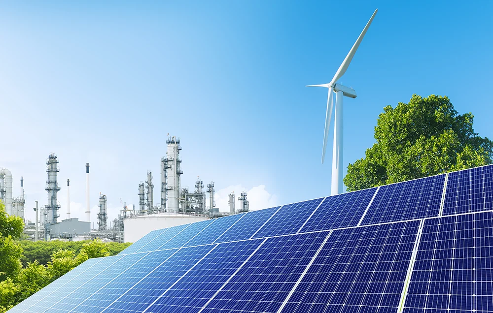 Solar panels in the foreground with a wind turbine and industrial plant in the background, illustrating a mix of renewable and traditional energy sources.