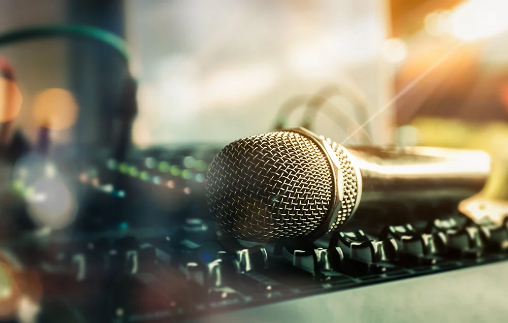 Close-up of a handheld microphone resting on an audio mixing console, with headphones and warm studio lighting in the background.