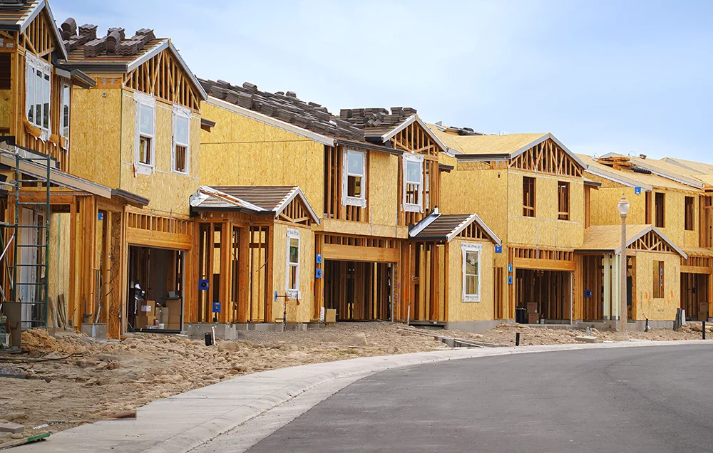 Row of newly constructed townhomes with exposed wooden framing and sheathing, unfinished driveways, and a curved residential street in front.