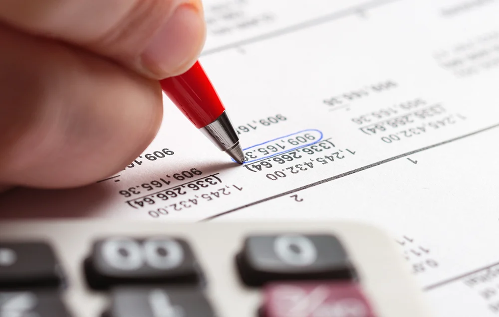 Close-up of a hand holding a red pen, pointing at and circling numbers on a printed financial or tax document, with a blurred calculator in the foreground.